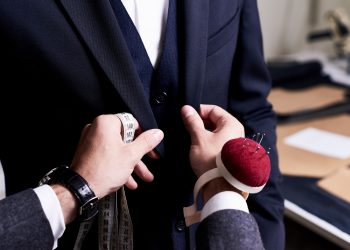 Closeup of tailor fitting bespoke suit to model, hands with tape measure and pin cushion fixing jacket on male model